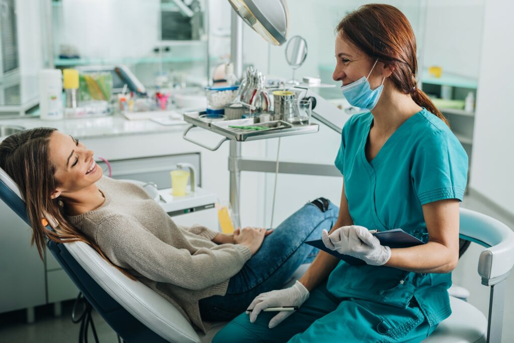A patient reclines in a dental chair wearing a beige sweater, while a dental professional in teal scrubs and white gloves stands beside them holding a clipboard. The clean clinical setting features organized dental tools and supplies, suggesting preparation for treatment or consultation.