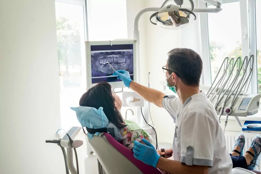 A dental professional points to a panoramic X-ray displayed on a monitor, discussing diagnostic details with a seated patient. The clinical setting includes gloves, protective gear, and dental equipment, emphasizing thorough evaluation and treatment planning.