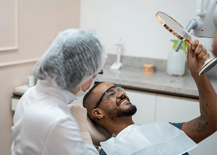 A patient reclines in a dental chair holding a mirror, while a dental professional in a white coat and hairnet performs an examination. Dental supplies on a nearby counter reinforce the clinical setting, highlighting patient engagement during treatment.