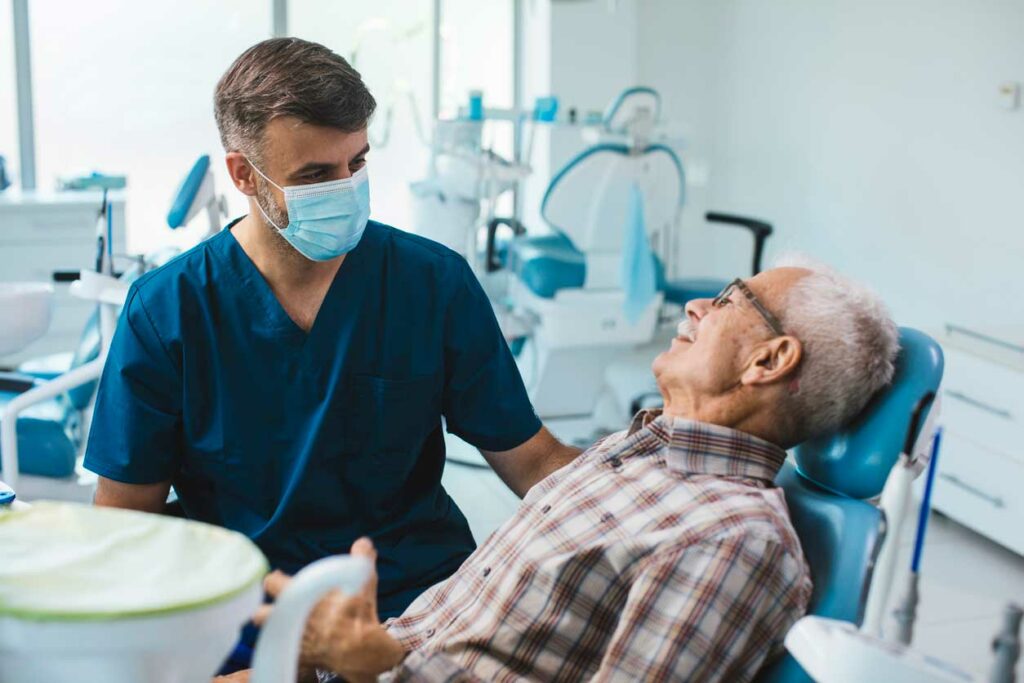 A dental professional in blue scrubs examines a seated patient wearing a plaid shirt inside a modern dental clinic. Equipment such as an overhead light, monitor, and instruments emphasize the clinical setting and active patient care.