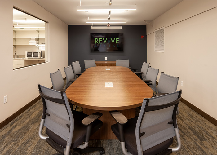 A conference room features a large wooden oval table surrounded by eight gray office chairs, with a wall-mounted screen displaying “REVIVE DENTAL.” Beige walls and patterned carpeting create a neutral backdrop, while a side window reveals a neighboring room containing shelves and kitchen appliances, contributing to the clinic’s multipurpose interior design.