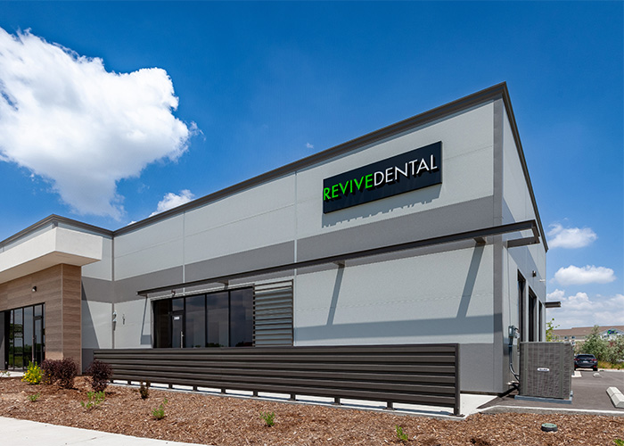 Modern dental clinic exterior with a 'REVIVE DENTAL' sign, featuring a sleek grey and white design, large window, and landscaped surroundings.