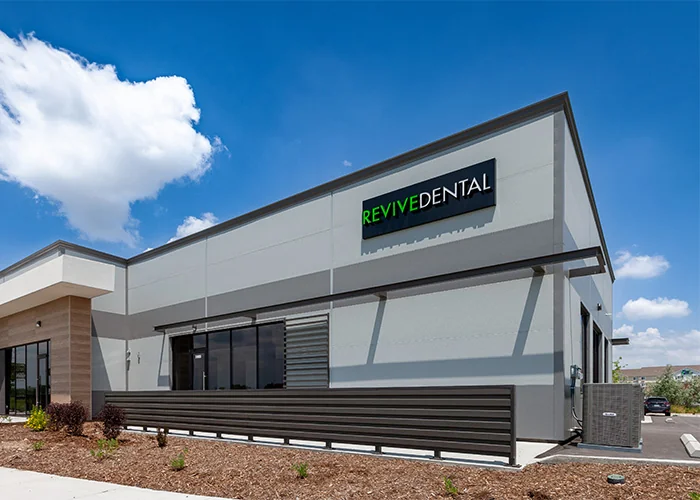 Modern dental clinic exterior with a 'REVIVE DENTAL' sign, featuring a sleek grey and white design, large window, and landscaped surroundings.