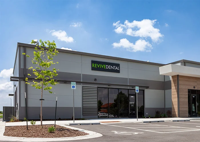 Modern dental clinic exterior with a 'REVIVE DENTAL' sign, featuring a sleek grey and beige design, reserved parking signs, and a landscaped area with a small tree under a clear sky.