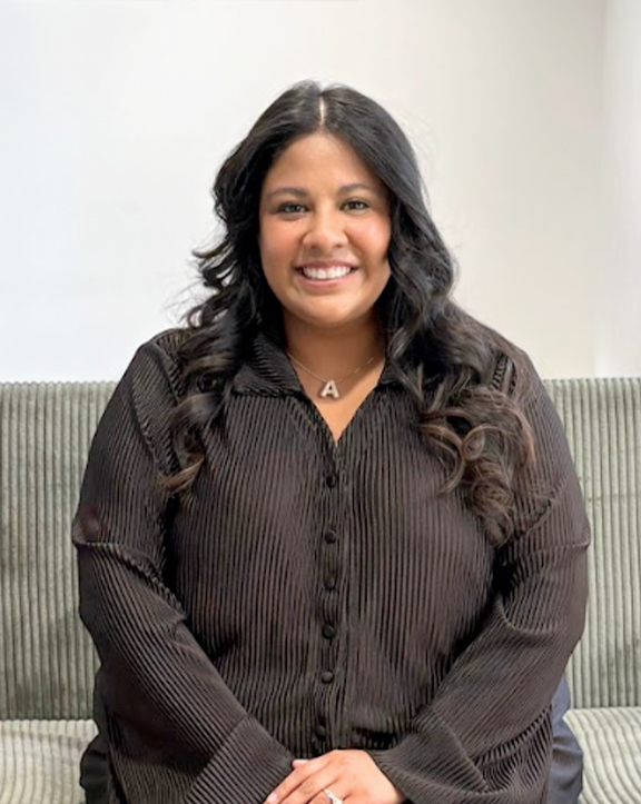 A person with long, wavy hair sits on a striped couch, wearing a black ribbed button-up top and a silver “A” pendant necklace. The light, plain background emphasizes their relaxed posture and polished, minimalist style. | Revive Dental implants and dentistry in Longmont, CO