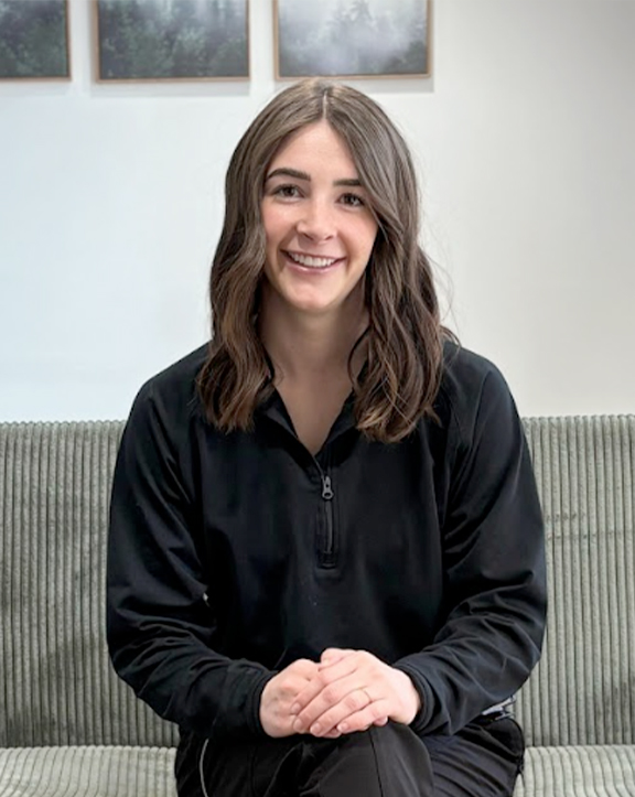 A person sits on a green couch wearing a black zip-collar long-sleeve shirt with hands clasped on their lap. Behind them, a light wall features three framed nature-themed pictures depicting trees and foliage, lending a serene, cozy atmosphere to the indoor setting. | Revive Dental implants and dentistry in Longmont, CO
