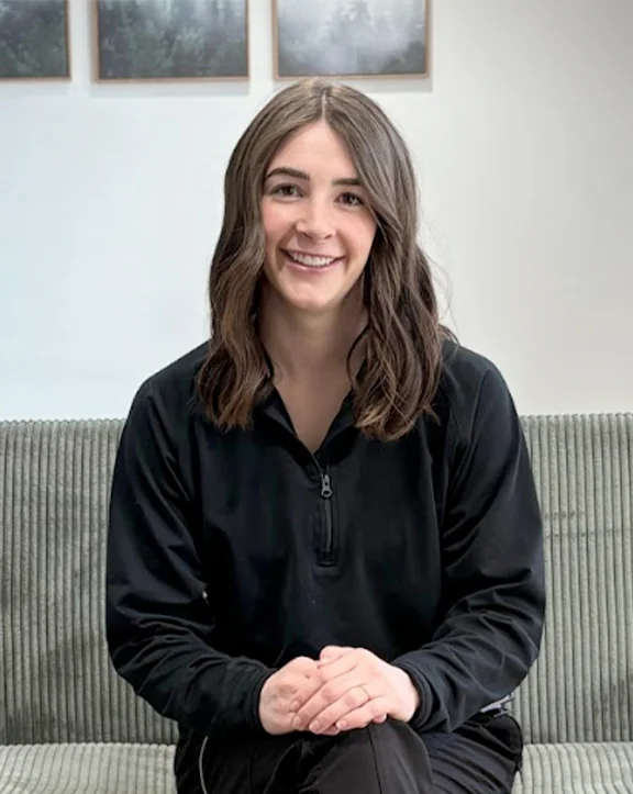 A person sits on a green couch wearing a black zip-collar long-sleeve shirt with hands clasped on their lap. Behind them, a light wall features three framed nature-themed pictures depicting trees and foliage, lending a serene, cozy atmosphere to the indoor setting. | Revive Dental implants and dentistry in Longmont, CO