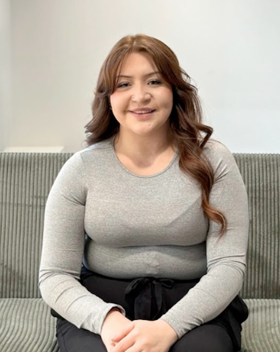 A person with long, reddish-brown wavy hair sits on a green couch, wearing a light gray long-sleeve top and black pants. The background is a plain, light-colored wall with no visible decor, emphasizing a simple and relaxed indoor setting. | Revive Dental implants and dentistry in Longmont, CO