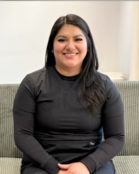 A person sits in a greenish-gray upholstered chair, layered in a black short-sleeve top over a black long-sleeve shirt. The setting appears to be indoors, with a white wall and partial window visible, presenting a simple, quiet moment in a minimally styled space. | Revive Dental implants and dentistry in Longmont, CO