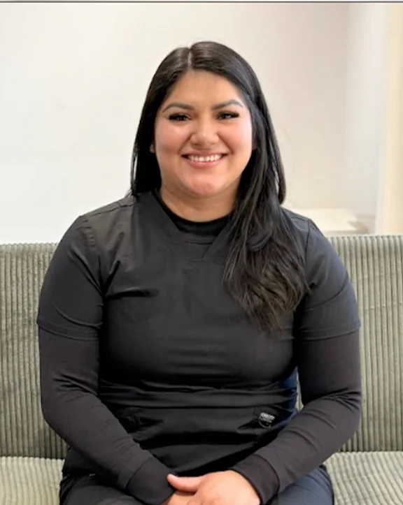 A person sits in a greenish-gray upholstered chair, layered in a black short-sleeve top over a black long-sleeve shirt. The setting appears to be indoors, with a white wall and partial window visible, presenting a simple, quiet moment in a minimally styled space. | Revive Dental implants and dentistry in Longmont, CO