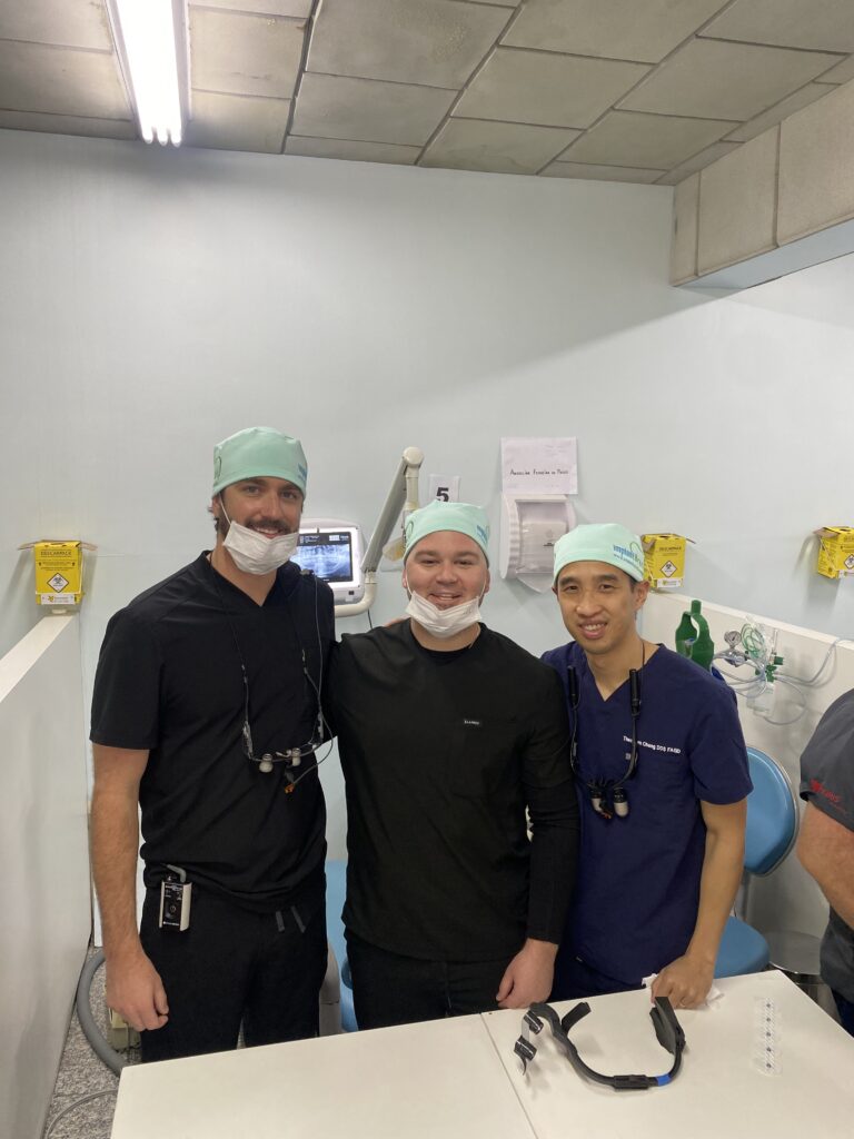 Three dental professionals stand together in a treatment room, wearing surgical masks and caps. Two are dressed in dark scrubs-one with dental loupes while the third wears a black shirt. Behind them are a monitor displaying a dental X-ray, a biohazard container, and signage in Spanish, reinforcing the clinical setting.
