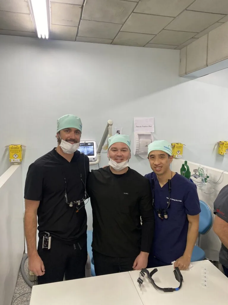 Three dental professionals stand together in a treatment room, wearing surgical masks and caps. Two are dressed in dark scrubs-one with dental loupes while the third wears a black shirt. Behind them are a monitor displaying a dental X-ray, a biohazard container, and signage in Spanish, reinforcing the clinical setting.
